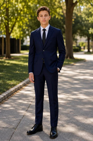 A young man wearing a navy blue suit, white dress shirt, and black tie, standing on a paved path in a park with trees in the background.