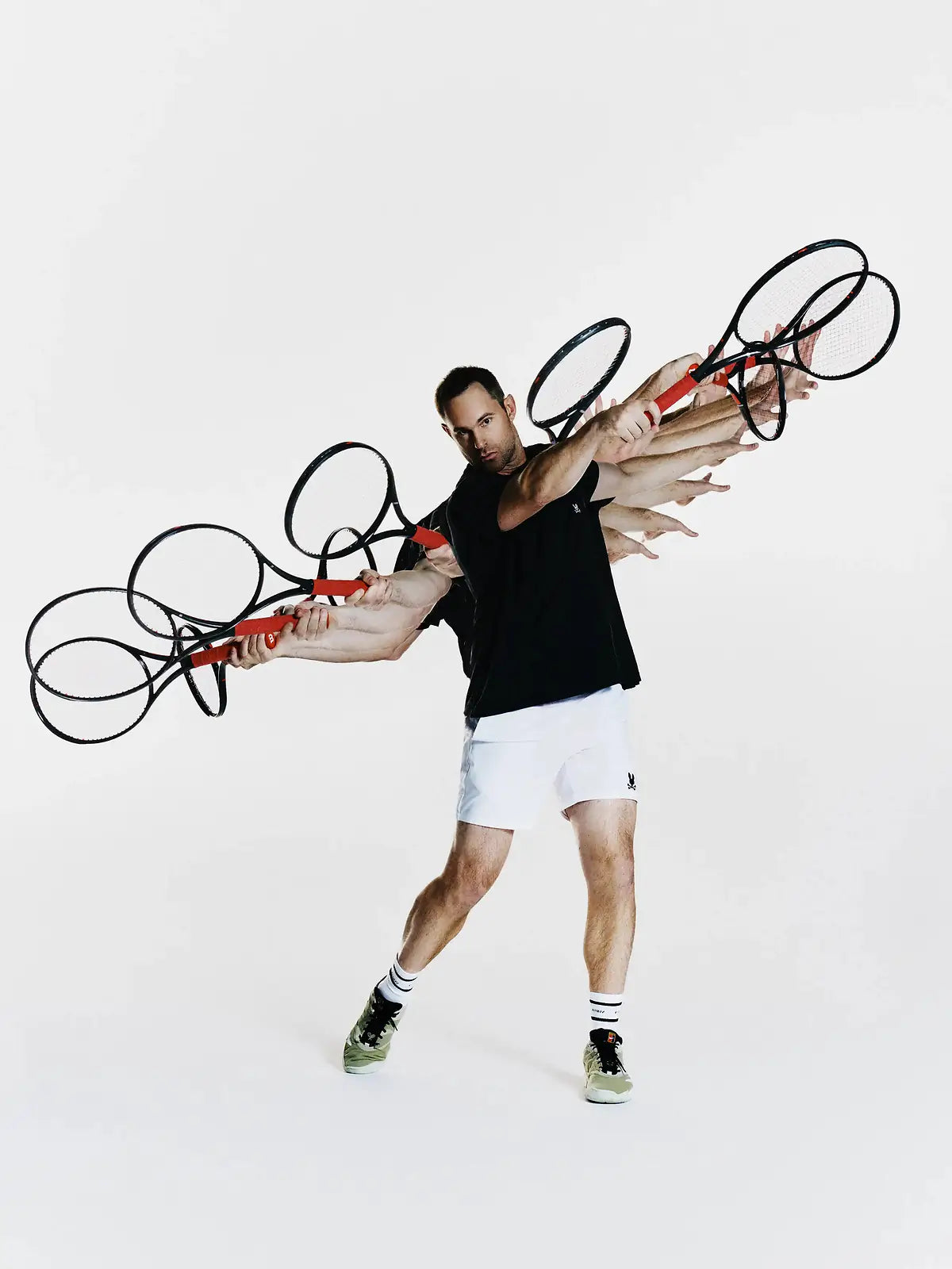 A man in a black tank top and white shorts poses dynamically while holding multiple tennis rackets with red handles and black frames, set against a plain white studio background.