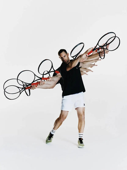 A man in a black tank top and white shorts poses dynamically while holding multiple tennis rackets with red handles and black frames, set against a plain white studio background.