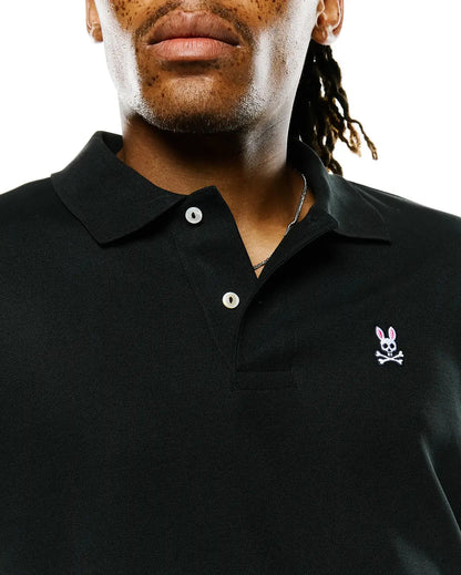 Close-up of a man wearing a black polo shirt with a small embroidered bunny skull and crossbones logo on the left chest, set against a plain white background.