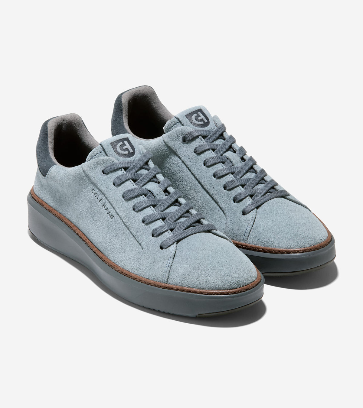 A pair of light blue suede low-top Cole Haan sneakers with dark gray laces, a thick dark gray sole, and a thin brown stripe detail on the side, shown against a light gray background.